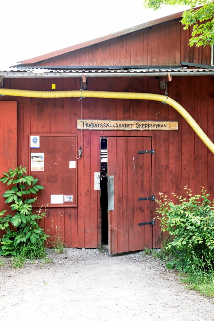 En dörr i en äldre falurödmålad byggnad står lite på glänt. Ovanför dörren står det "Träbåtssällskapet Skeppsmyran" på en träskylt.