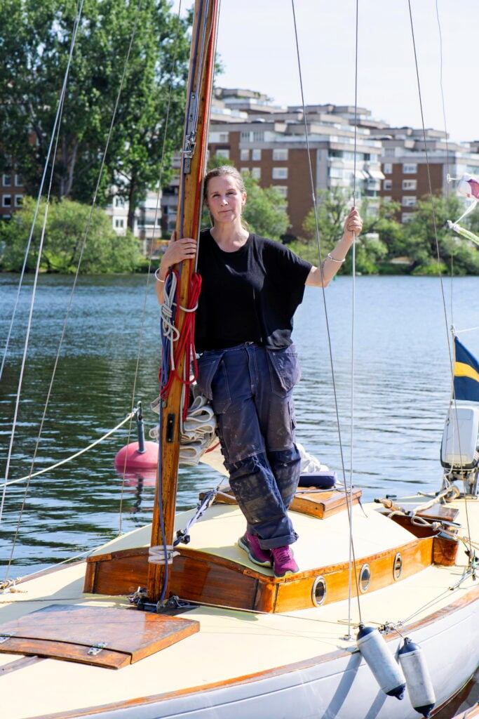 En kvinna iförd snickarbyxor och t-shirt står vid masten på en äldre träsegelbåt.