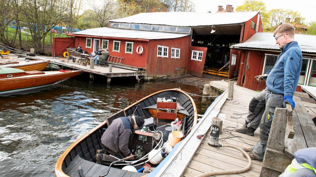 En man i en snipa på knä i båten. På bryggan står en man och pekar.