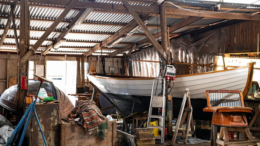 En stökig interiör i en låg träbyggnad med glest mellan brädorna. En snipa med vitt skrov till höger i bild och en uppochnedvänd träbåt till vänster.