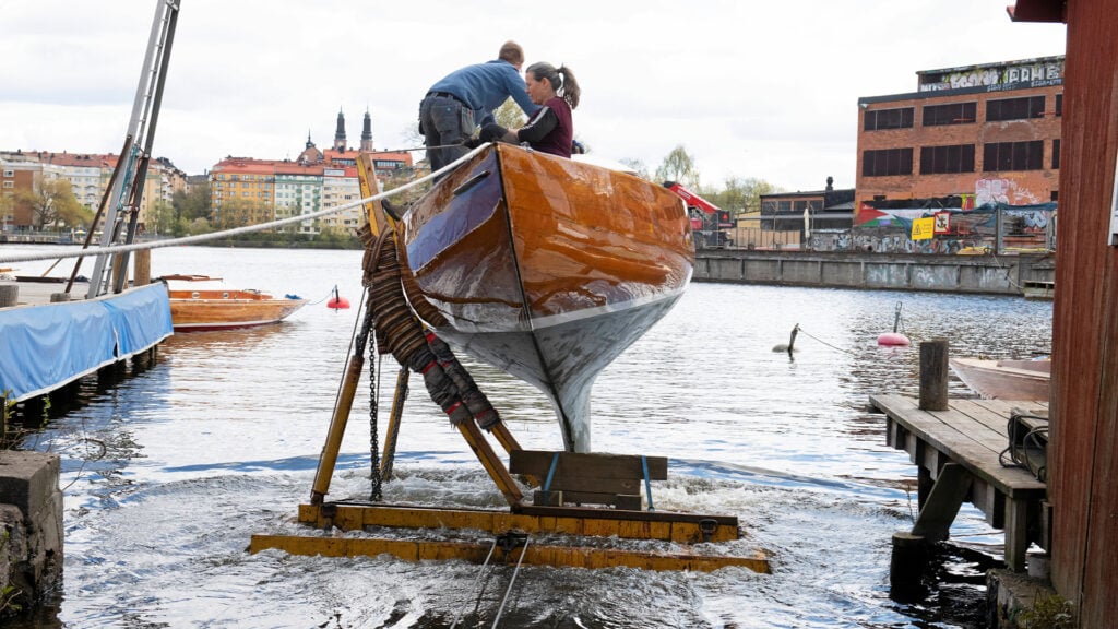 En man och en kvinna i en omastad träsegelbåt med blankt skrov, som är på väg ner i vattnet på en slip.
