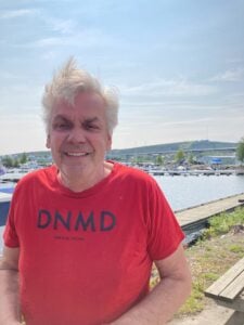 En leende man iförd röd tröja, framför en småbåtshamn.