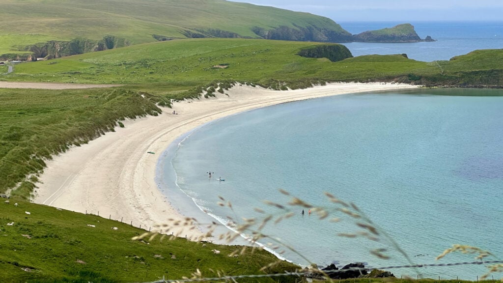 Bild tagen ovanifrån över vacker sandstrand i en halvmåne till vänster i bild. Man anar några badande och två kanotister.