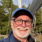 En man med keps, glasögon och kort, vitt skägg.