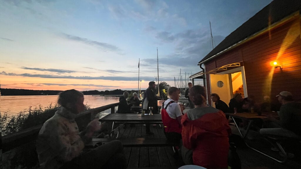 Ekolns batklubb i kvallsljus. Ett antal personer sitter i kvällningen utanför ett litet hus vid vattnet.
