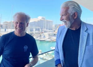 Två män på en balkong med hav och en småbåtshamn i bakgrunden.