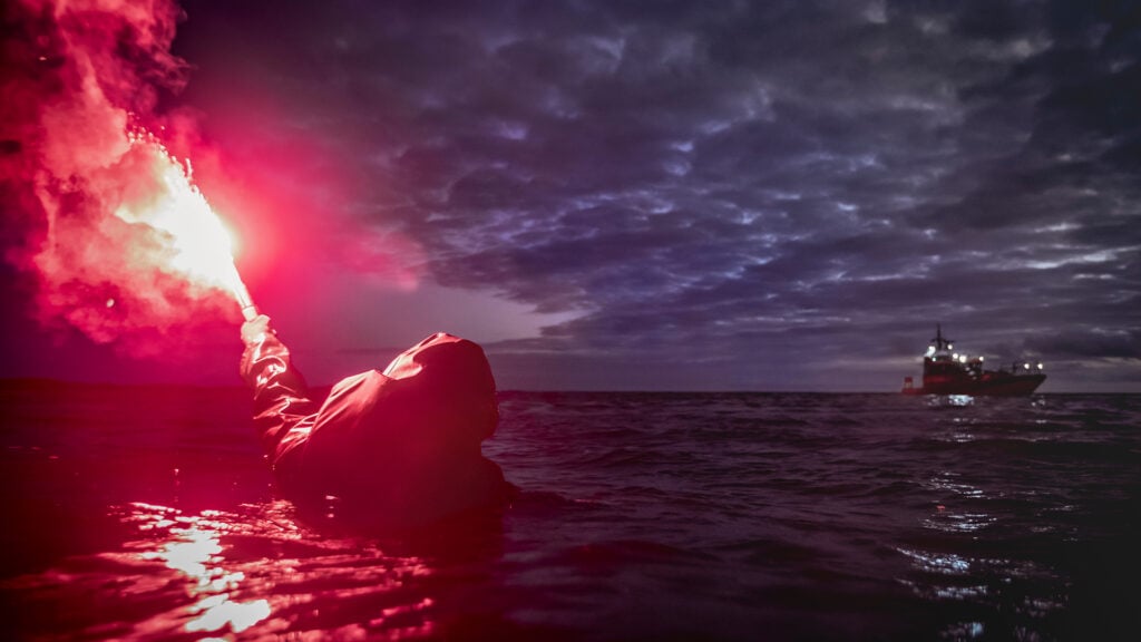 En person ligger i vattnet till havs med ett nödbloss i handen. I bakgrunden en av SSRS räddningsbåtar.